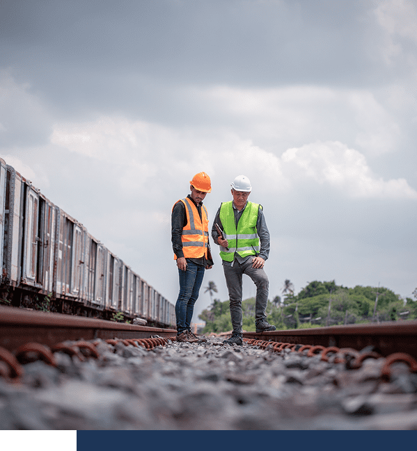 Two workers wearing safety vests and helmets inspect railroad tracks outdoors, with train cars visible on the left and cloudy sky overhead.